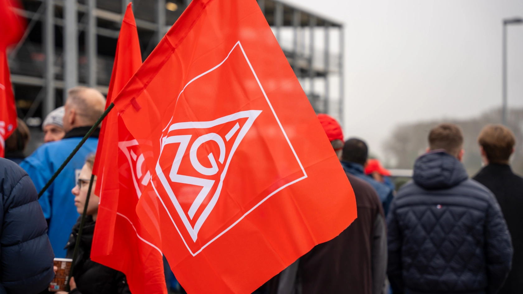 IG Metall-Fahne - im Hintergrund Beschäftigte bei einem Warnstreik 