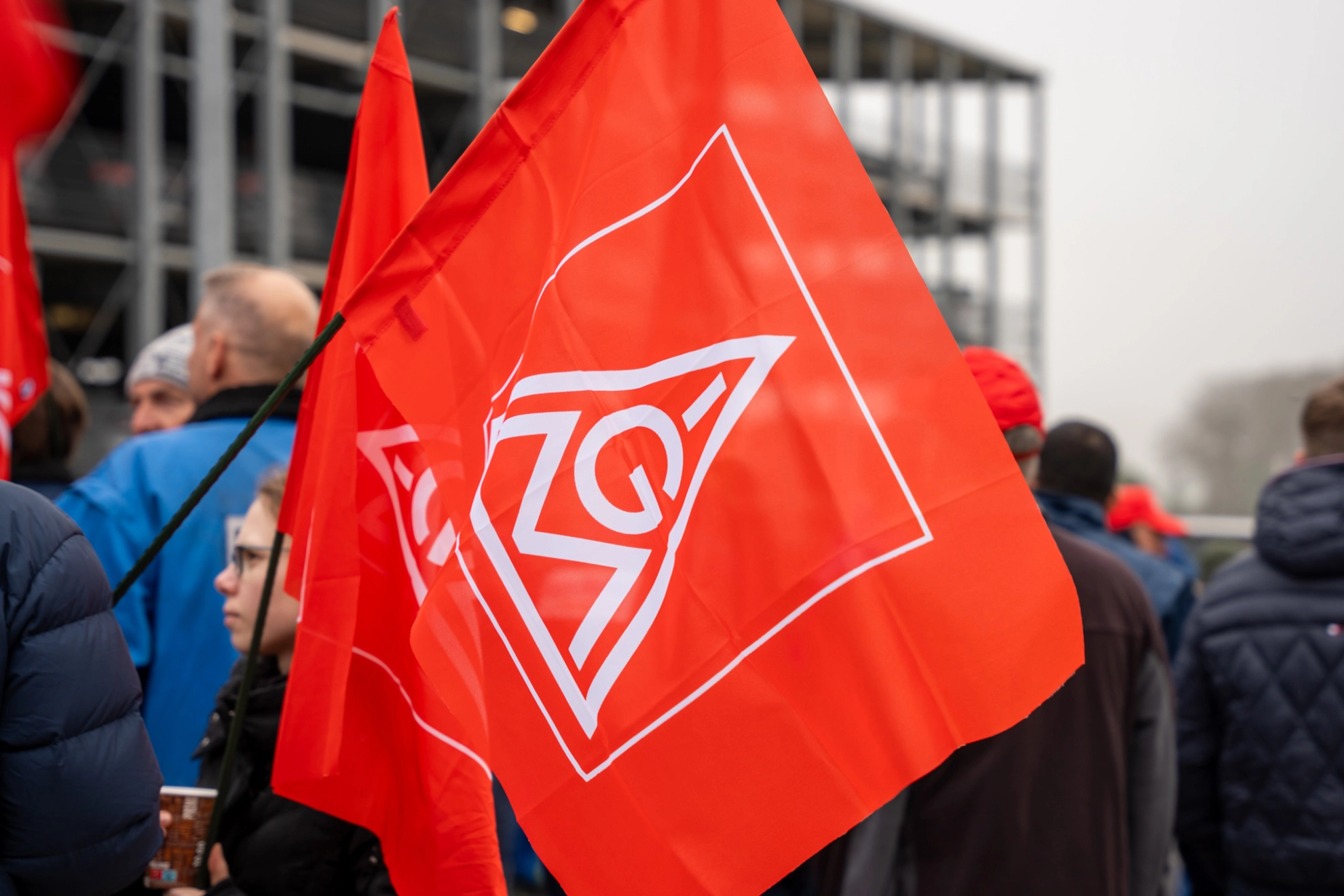 IG Metall-Fahne - im Hintergrund Beschäftigte bei einem Warnstreik 